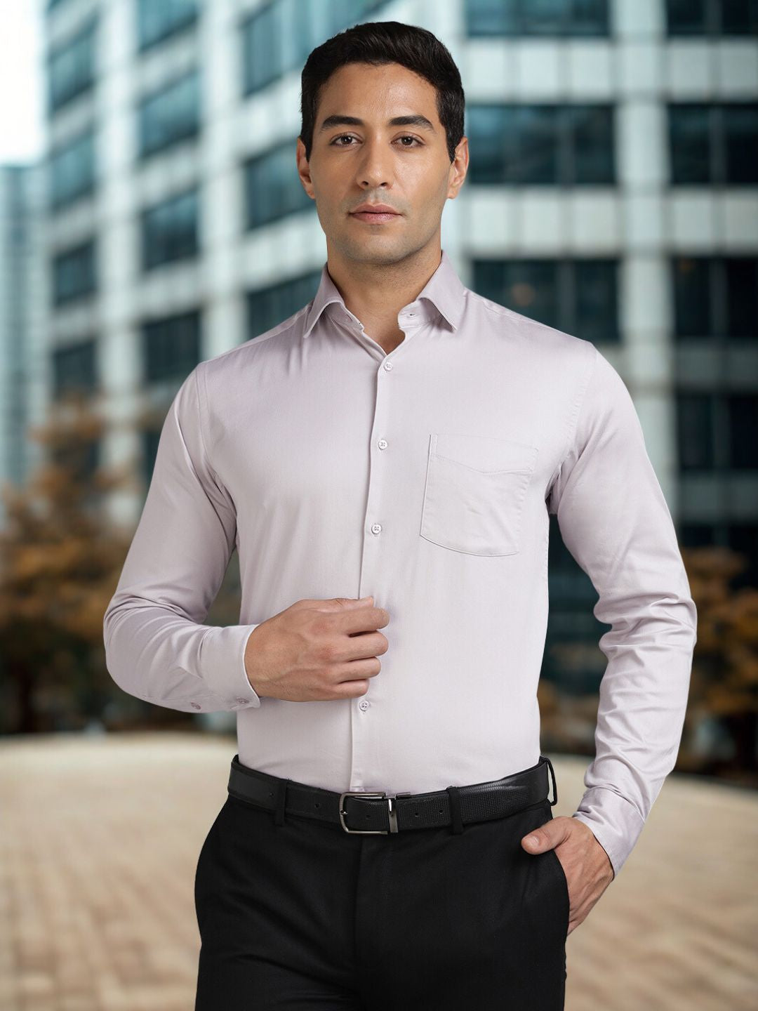 Man wearing a light grey dress shirt and black pants standing in front of a blurred cityscape.