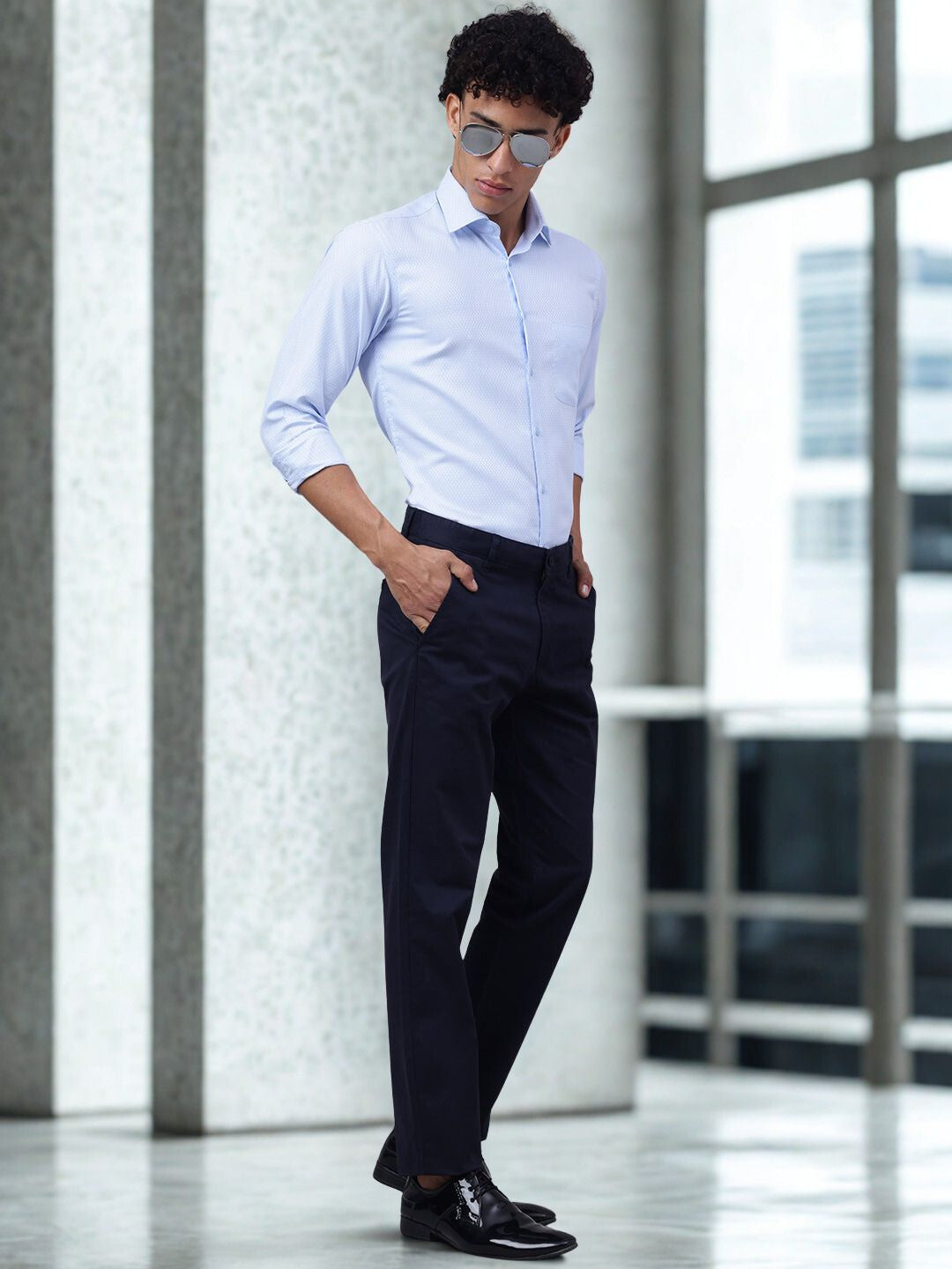 Man wearing a light blue shirt and dark pants in an indoor setting with large windows.