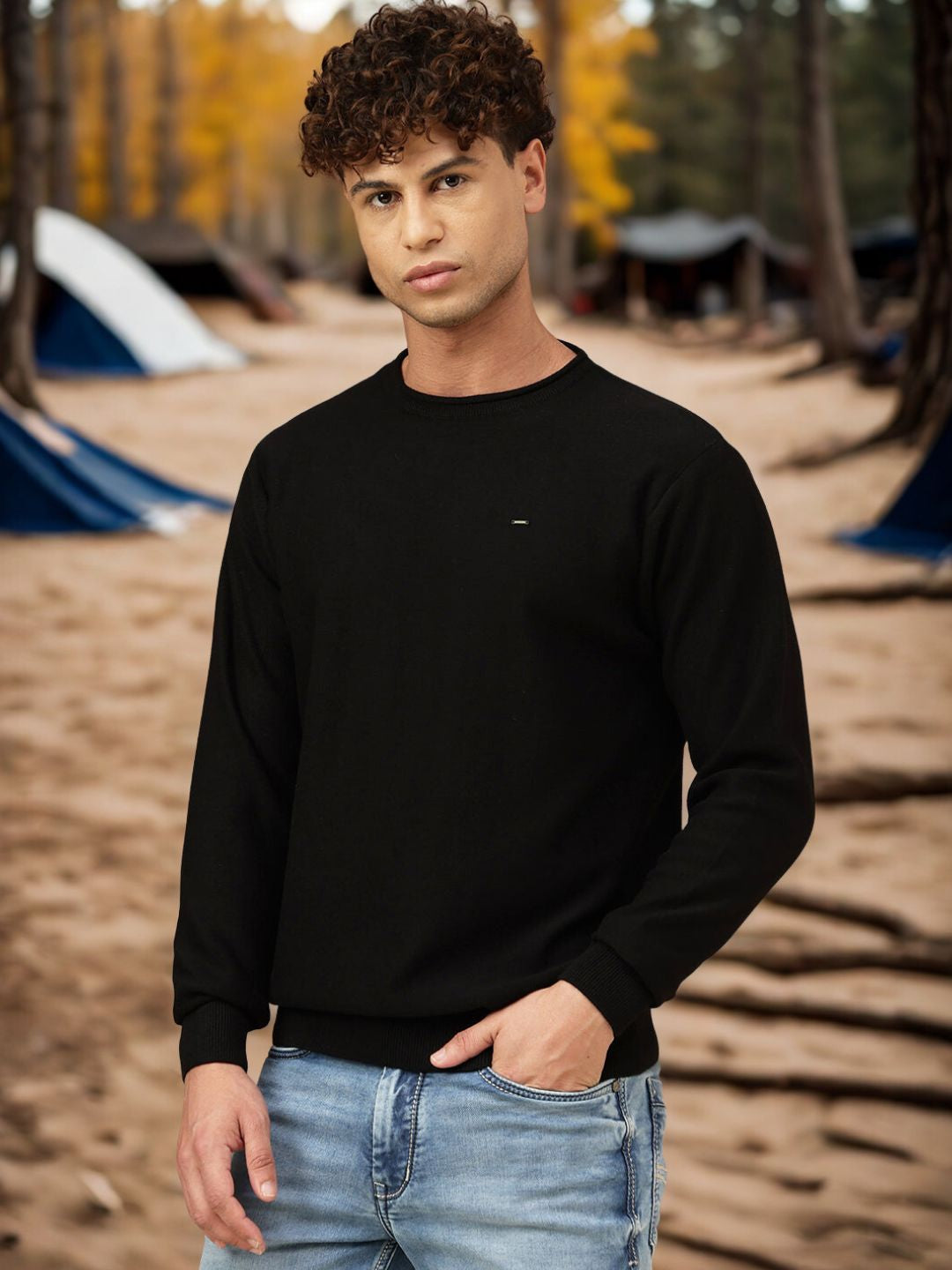 Person wearing a black sweater and jeans standing in a forest with camping tents in the background
