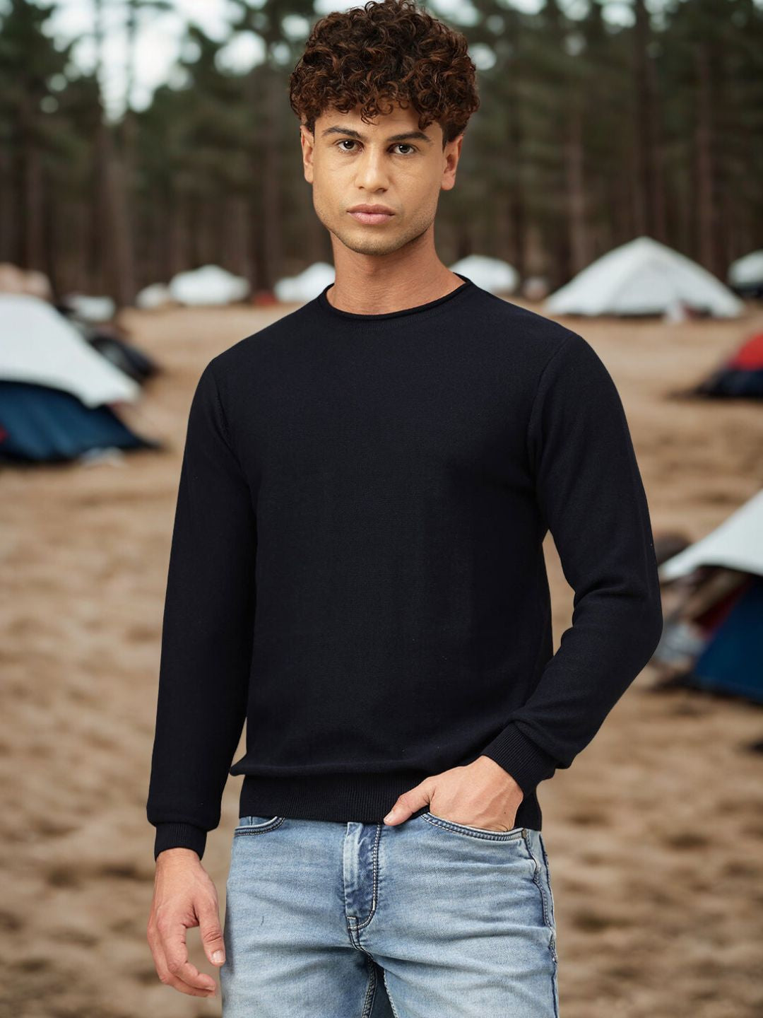 Man wearing a black sweater and light blue jeans standing in a camping area with tents in the background.