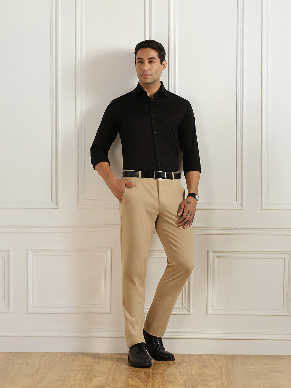 Man wearing a black shirt and beige pants standing against a white paneled wall.