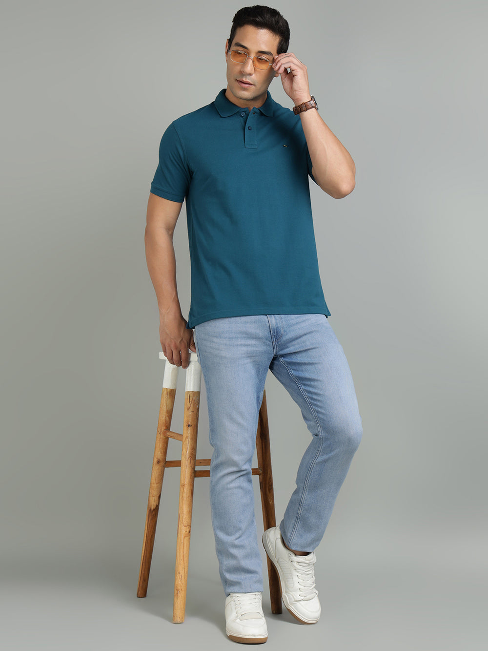 Man wearing a teal polo shirt and light blue jeans, standing on a wooden stool against a gray background.