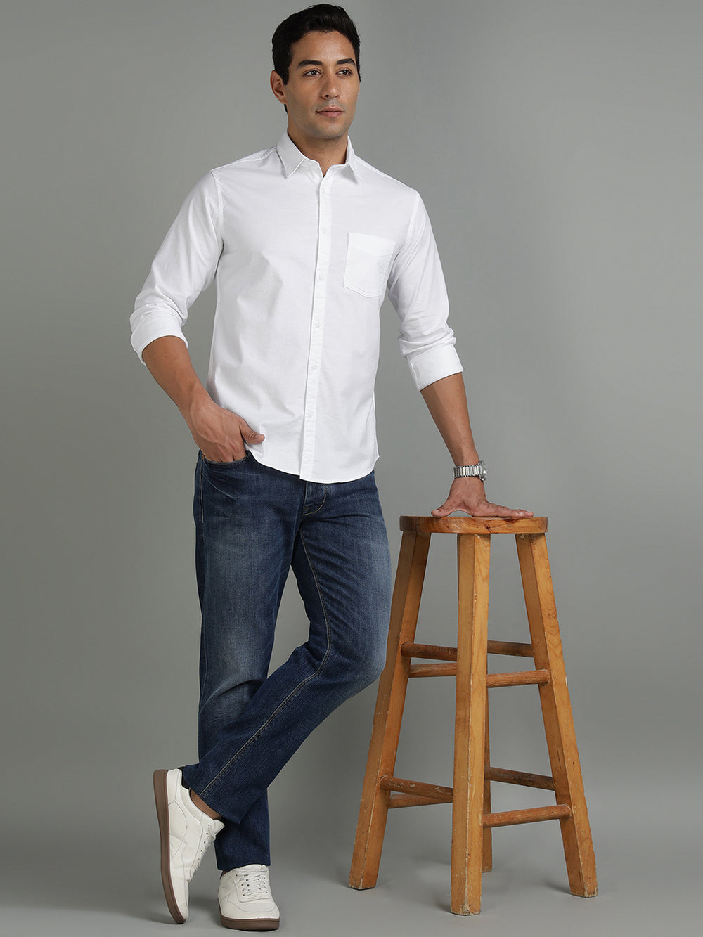Man wearing a white shirt and blue jeans standing next to a wooden stool on a gray background