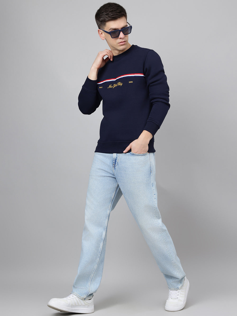 Man wearing a navy sweater with a logo, light blue jeans, and white sneakers on a gray background